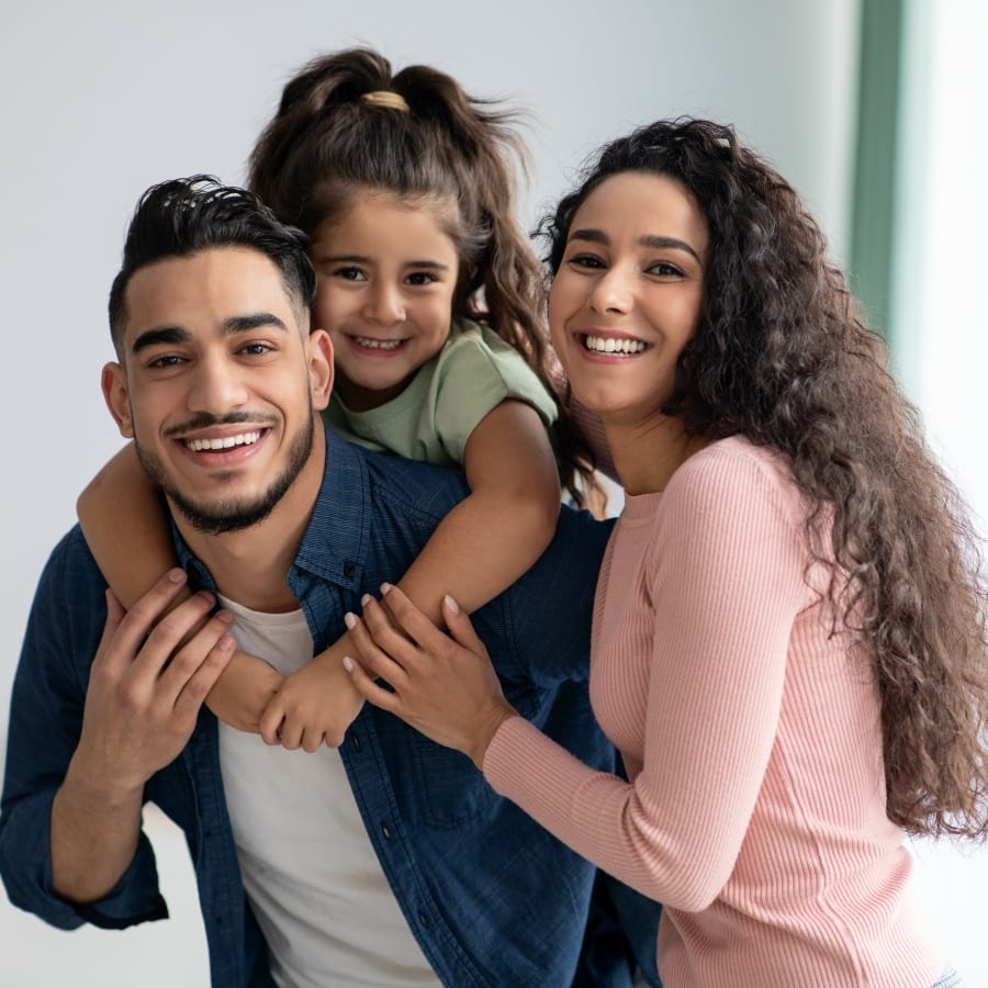 A young girl is riding piggyback on a man, presumably her father, as a woman, presumably her mother, hugs them both.