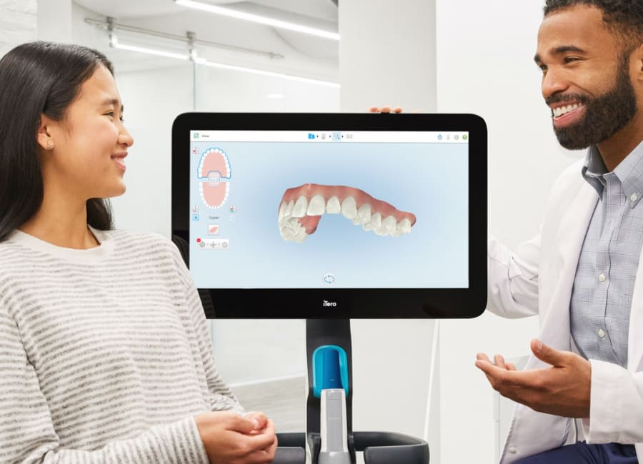 A dentist and a patient discuss oral health. A digital rendering of the patient's teeth is on the screen behind them.