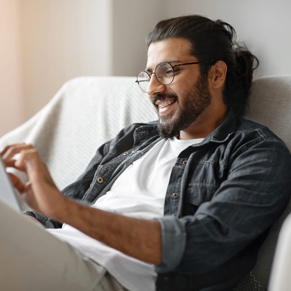 A man with long hair and a beard is smiling at a tablet. He is wearing circular glasses and has braces.